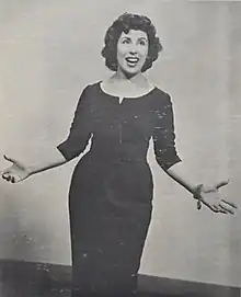 A young woman with fair skin and dark hair, standing with forearms out to her sides, singing