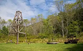 A wooden derrick at left on grass, with trees and a mountain ridge in the background