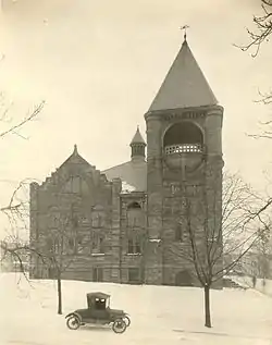 A stone building with tall windows and a pyramidal tower on the right, with snow on the ground and an automobile passing in front