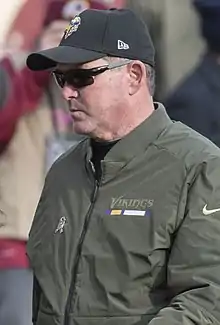 Color photograph of Zimmer walking on a football field, wearing a green jacket and black baseball cap with the Minnesota Vikings logo.