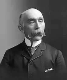 Portrait of Peter Bryce. Wearing a jacket and tie, he is looking off-camera with an expressionless face