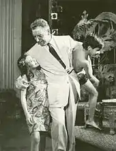 Pinza, in a white suit, walks along holding a mixed-race boy, about 7 or 8 years old, while listening to a slightly older girl who walks next to him.