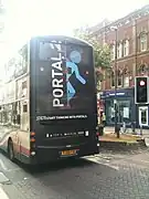 The back of a brown, double-decker bus in a city street, with the text "Portal 2" and a blue symbol of a man that seems to be vanishing through a doorway