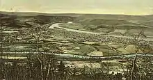 A tinted postcard depicts a town as seen from a hillside. It is laid out on a triangle of flat land between two converging streams. The larger and more distant of the two streams is flowing from mountains, while the smaller stream flows through farmland.