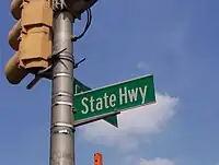 A green street sign reading State Highway attached to a traffic light pole.