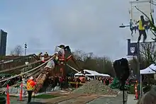 Action shot of people, wearing orange and yellow construction clothing, working to raise the Reconciliation Pole at UBC