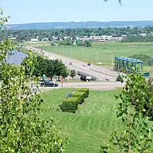 A concrete and steel structure holds a mildly busy highway crossing the river seen to the left. There are marshes and a city in the background.