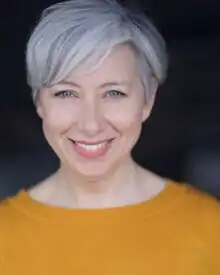 White woman with grey hair, wearing a yellow pullover jumper, smiling at the camera