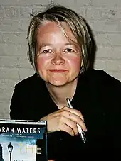 Image of a woman with short blond hair seated at a table holding a pen and a book cover to the camera