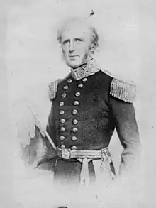 Picture from waist up of a standing pale-skinned man, balding, with bushy side-burns, wearing double-breasted dark uniform with heavy gold epaulettes, gold braid, and a sash