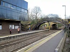 A station with two platforms and tracks. In the foreground is a 19th-century pedestrian bridge.