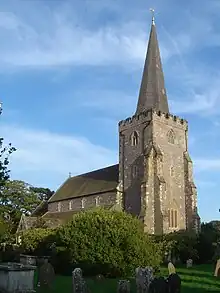 Three-quarter view of a stone church with a buttressed tower in the foreground. This has small battlements and a spire. The nave roof, below which are four small, evenly spaced windows, is visible, but its aisle and an attached porch are obscured by a bush. There are gravestones and a table tomb in the foreground.