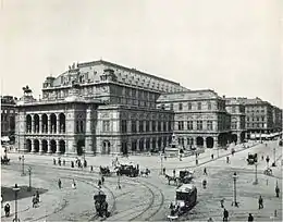 An imposing, heavily ornamented building in a city location, with numerous horsedrawn vehicles and pedestrians passing. There are visible tramlines in the street.