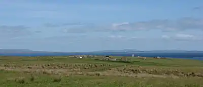 View looking north showing grass fields in the foreground, with ruined buildings visible in the middle distance and sea and islands visible on the horizon