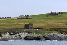 View of ruined houses and a ruined building surrounded by a stone wall, located on a green slope leading down to a rocky shoreline