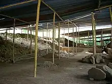 A cleared area of bare soil strewn with rubble, with raised areas to the left and right and roofed with corrugated metal supported by timber poles