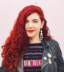 A young woman with curly red hair wearing a jacket stands in front of a pink-painted brick wall with her arms partially outstretched and her palms facing upward.