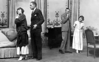 Stage scene, with middle-aged white man and women to left, in discussion, and two younger adults tiptoeing away