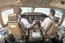 A flight deck, from behind the two pilots' seats. A center console lies in between the seats, in front is an instrument panel with several displays, and light enters through the forward windows.