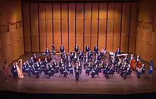 Picture of the United States Air Force Concert Band. The members of the music ensemble are seated in a semi-circular formation of rows on a stage wearing the dark blue Air Force mess dress uniform and holding their instruments. The conductor of the band in stand in the center of the picture.