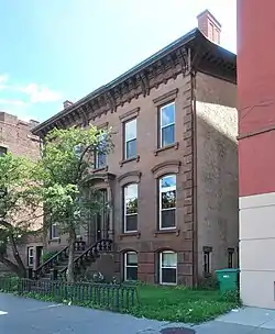 An ornate brown stone building three stories high with a flat roof and small yard in front on a city street