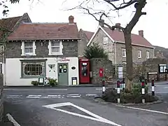 The markings on a road junction around a tree are visible in the foreground, in front of a small shop with Christmas decorations in the window. A red K8 model telephone box and a red post box built into a wall are to the left.