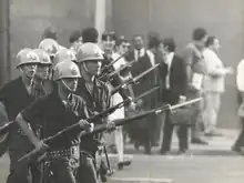 Several men in a line wear helmets and brandish rifles with bayonets